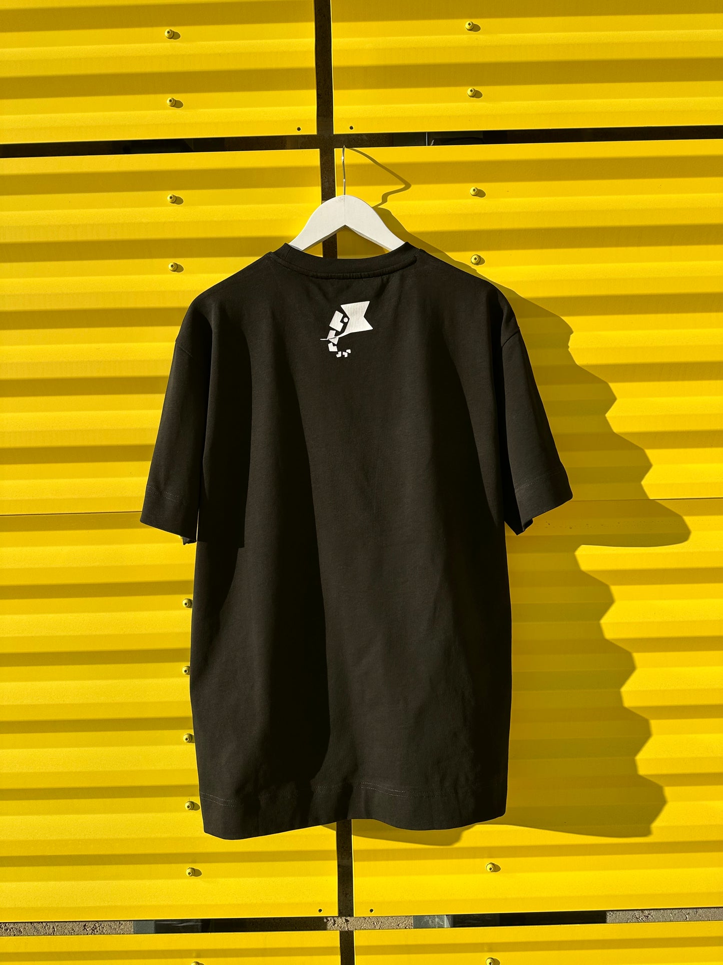 A black everyday T-shirt displayed on a hanger against a yellow background, with a small white logo on the back.