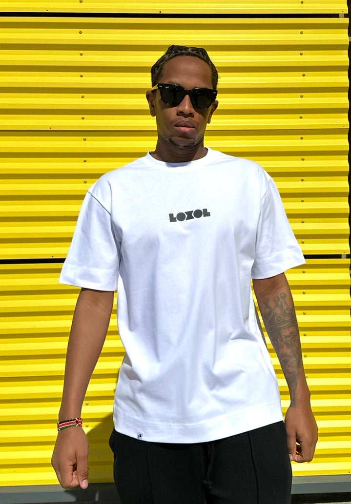 A man wearing a white crewneck t-shirt with 'LOXOL' printed in black on the chest, paired with black pants and sunglasses, standing in front of a bright yellow corrugated metal wall.